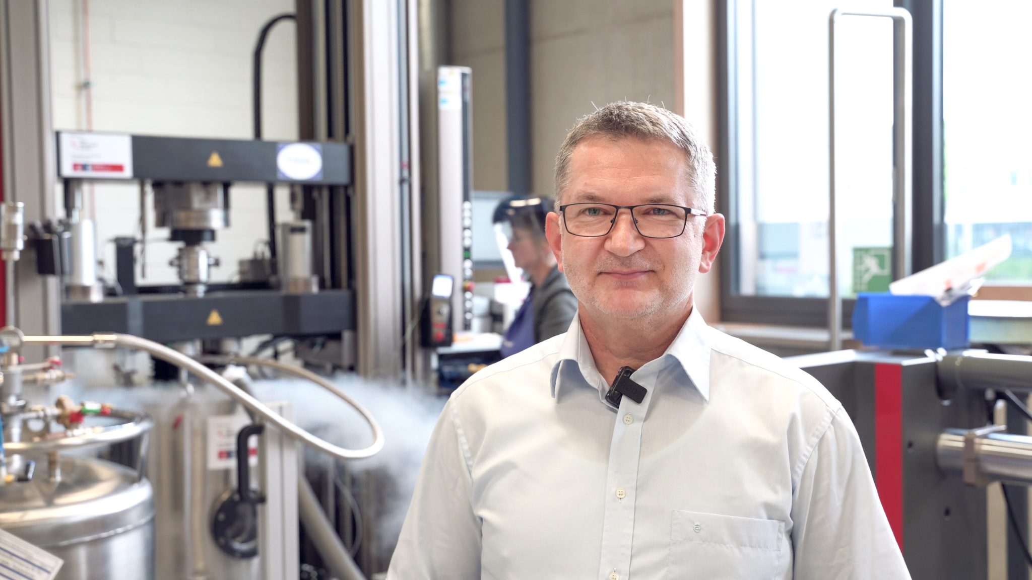 Ein Mann mit Brille und hellem Hemd steht lächelnd in einem modernen Labor oder technischen Prüfraum. Im Hintergrund sind technische Geräte, Rohre und eine Arbeitskraft mit Schutzvisier zu sehen. Es steigt sichtbar Dampf aus einem Edelstahlbehälter auf. Das Bild vermittelt einen Einblick in ein hochtechnologisches Arbeitsumfeld.