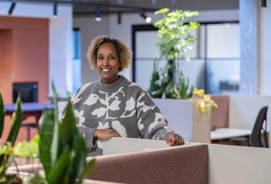Eine lächelnde Frau steht in einem modernen, freundlich gestalteten Büro mit vielen Pflanzen. Sie trägt einen grauen Pullover mit weißem Blumenmuster und lehnt sich locker an eine halbhohe Trennwand. Im Hintergrund sind Arbeitsplätze mit Bürostühlen und weiteren Pflanzen zu sehen.