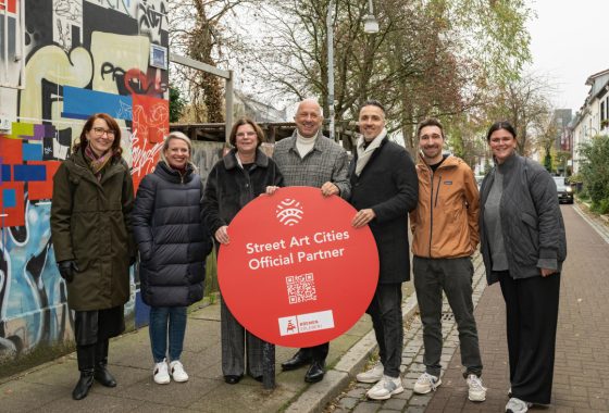 Acht Personen stehen lächelnd auf einem Gehweg in einer Wohnstraße vor einer bunt besprayten Wand. In ihrer Mitte halten sie ein großes, rundes rotes Schild mit der Aufschrift „Street Art Cities Official Partner“. Darunter ist ein QR-Code sowie das Logo von „Bremen Erleben“ zu sehen. Die Gruppe ist warm gekleidet, es ist Herbst oder Winter.