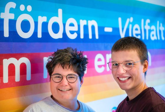Zwei fröhlich lächelnde Personen mit kurzen Haaren und Brillen stehen nebeneinander vor einem farbenfrohen Hintergrund mit Regenbogenfarben und großem Text. Der sichtbare Teil des Textes lautet „fördern – Vielfalt“. Das Bild vermittelt Offenheit, Vielfalt und Zusammenhalt.