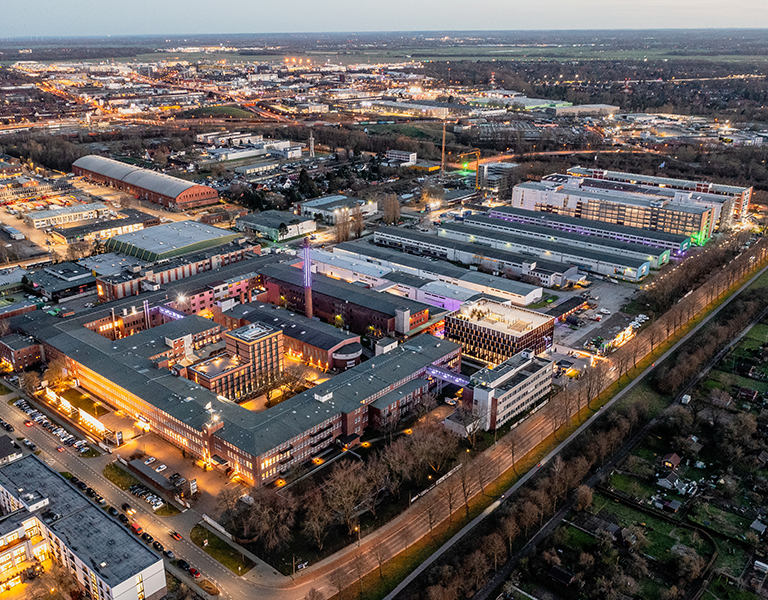 Luftaufnahme des Gewerbe- und Kreativquartiers "Tabakquartier" in Bremen bei einsetzender Dämmerung. Die Gebäude sind stimmungsvoll beleuchtet, insbesondere der zentrale Gebäudekomplex mit dem markanten Schornstein. Umgeben ist das Areal von weiteren Hallen, Büros und Wohngebäuden, im Hintergrund sind Wohnsiedlungen und grüne Flächen zu erkennen. Die Straßen und Wege sind beleuchtet, die Szene wirkt lebendig und urban.
