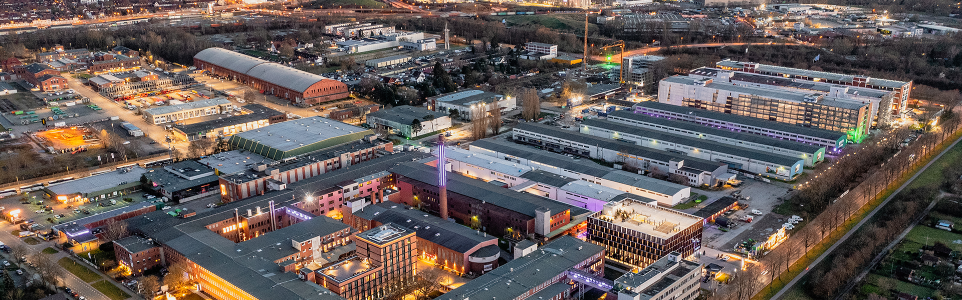 Luftaufnahme des Gewerbe- und Kreativquartiers "Tabakquartier" in Bremen bei einsetzender Dämmerung. Die Gebäude sind stimmungsvoll beleuchtet, insbesondere der zentrale Gebäudekomplex mit dem markanten Schornstein. Umgeben ist das Areal von weiteren Hallen, Büros und Wohngebäuden, im Hintergrund sind Wohnsiedlungen und grüne Flächen zu erkennen. Die Straßen und Wege sind beleuchtet, die Szene wirkt lebendig und urban.