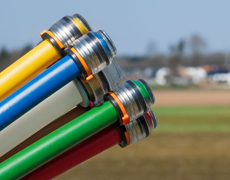 Nahaufnahme von mehreren farbigen Glasfaserleitungen mit Metallanschlüssen. Die Leitungen sind in Gelb, Blau, Weiß, Grün, Rot und Braun eingefärbt und zeigen in den Himmel. Im Hintergrund ist unscharf eine ländliche Landschaft mit Feldern und einigen Häusern zu sehen.