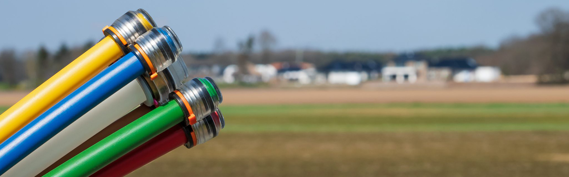 Nahaufnahme von mehreren farbigen Glasfaserleitungen mit Metallanschlüssen. Die Leitungen sind in Gelb, Blau, Weiß, Grün, Rot und Braun eingefärbt und zeigen in den Himmel. Im Hintergrund ist unscharf eine ländliche Landschaft mit Feldern und einigen Häusern zu sehen.