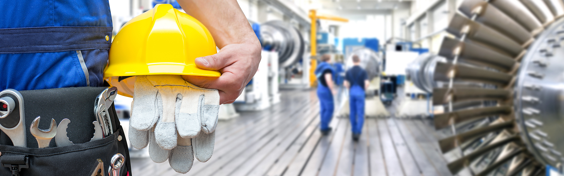 Ein Industriearbeiter in blauer Arbeitskleidung hält einen gelben Schutzhelm und ein Paar Arbeitshandschuhe in der Hand. Am Gürtel trägt er diverses Werkzeug. Im Hintergrund ist eine große, helle Werkhalle mit weiteren Personen in blauer Arbeitskleidung zu sehen.