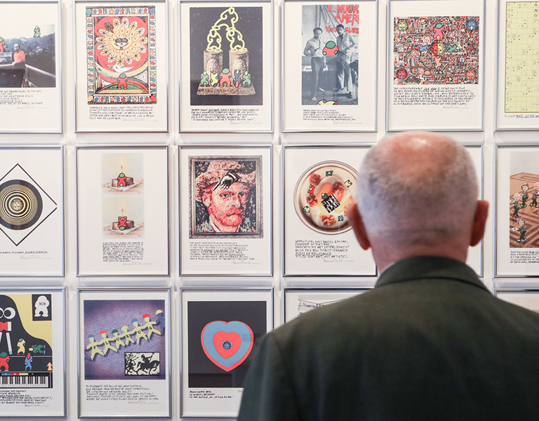 Ein älterer Mann mit Glatze und grünem Sakko betrachtet eine Wand mit gerahmten Kunstwerken. Die Bilder sind vielfältig gestaltet – darunter Zeichnungen, Collagen und Fotografien mit bunten Motiven und Texten. Die Szene spielt in einer Galerie oder Ausstellung.