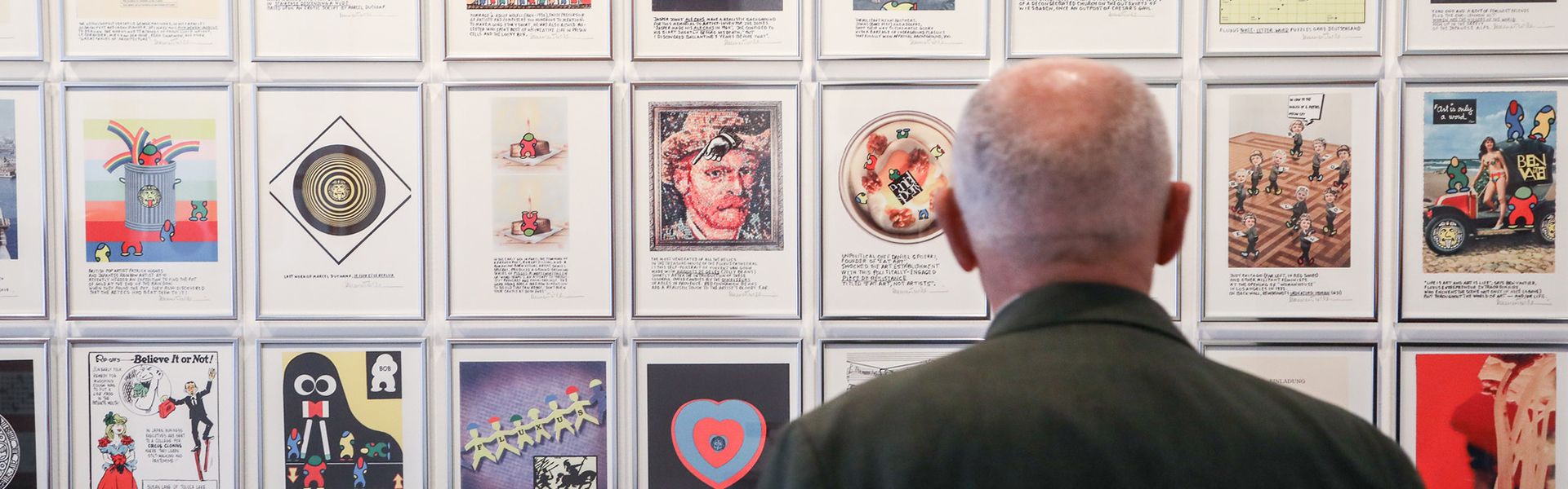 Ein älterer Mann mit Glatze und grünem Sakko betrachtet eine Wand mit gerahmten Kunstwerken. Die Bilder sind vielfältig gestaltet – darunter Zeichnungen, Collagen und Fotografien mit bunten Motiven und Texten. Die Szene spielt in einer Galerie oder Ausstellung.