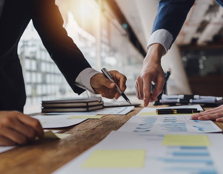 Mehrere Personen in Businesskleidung beugen sich über einen Tisch, auf dem Dokumente mit Diagrammen, Notizen und Stiften verteilt sind. Hände zeigen auf bestimmte Stellen in den Papieren – es scheint sich um eine konzentrierte Besprechung oder gemeinsame Analyse von Geschäftszahlen zu handeln. Im Hintergrund ist ein modernes Büro mit viel Tageslicht zu sehen.