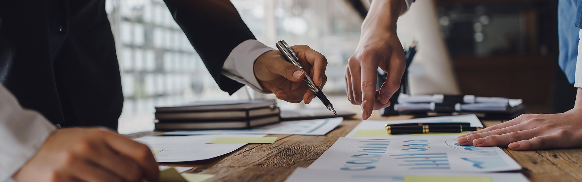 Mehrere Personen in Businesskleidung beugen sich über einen Tisch, auf dem Dokumente mit Diagrammen, Notizen und Stiften verteilt sind. Hände zeigen auf bestimmte Stellen in den Papieren – es scheint sich um eine konzentrierte Besprechung oder gemeinsame Analyse von Geschäftszahlen zu handeln. Im Hintergrund ist ein modernes Büro mit viel Tageslicht zu sehen.