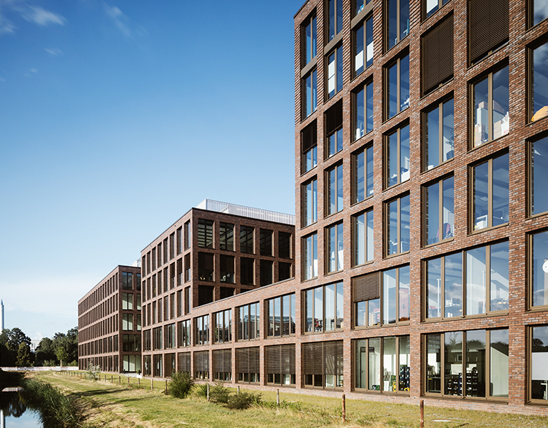 Moderne Bürogebäude aus rotem Backstein mit großen Glasfassaden erstrecken sich entlang eines begrünten Wasserlaufs in einem urbanen Umfeld. Die Architektur ist geradlinig und offen gestaltet, mit klaren Strukturen und großzügigen Fensterfronten, die viel Tageslicht ins Innere lassen. Der Himmel ist klar und blau – eine ruhige, professionelle Atmosphäre entsteht.