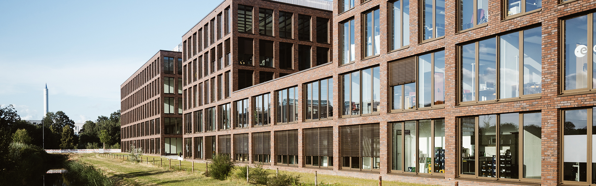 Moderne Bürogebäude aus rotem Backstein mit großen Glasfassaden erstrecken sich entlang eines begrünten Wasserlaufs in einem urbanen Umfeld. Die Architektur ist geradlinig und offen gestaltet, mit klaren Strukturen und großzügigen Fensterfronten, die viel Tageslicht ins Innere lassen. Der Himmel ist klar und blau – eine ruhige, professionelle Atmosphäre entsteht.