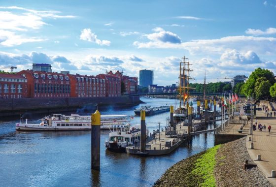 Panoramablick auf die Bremer Schlachte bei Sonnenschein. Mehrere Ausflugs- und Segelschiffe liegen am Steg, darunter ein großes Segelschiff mit gelbem Mast. Am rechten Bildrand verläuft eine belebte Uferpromenade mit Menschen, Cafés und Fahnen. Auf der linken Seite prägen rote Backsteingebäude das Uferbild, im Hintergrund ist ein moderner Glasbau zu sehen. Der Himmel ist blau mit einigen weißen Wolken.
