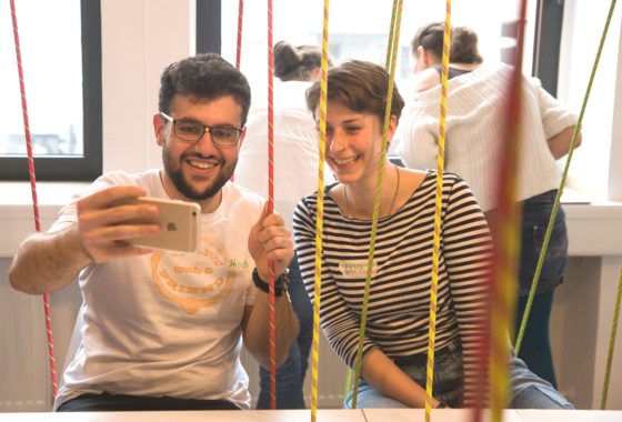 Ein junger Mann und eine junge Frau sitzen nebeneinander an einem Tisch und machen gemeinsam ein Selfie. Zwischen ihnen hängen bunte Kletterseile von der Decke. Beide lachen und wirken fröhlich. Im Hintergrund sind weitere Personen an Tischen beschäftigt. Die Atmosphäre ist locker und kreativ.