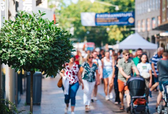 Im Vordergrund ist ein rund geschnittener, grüner Straßenbaum vor einem Gebäude zu sehen. Im Hintergrund läuft eine Menschenmenge durch eine sonnige Einkaufsstraße mit Ständen und Läden. Die Szene wirkt belebt und sommerlich.