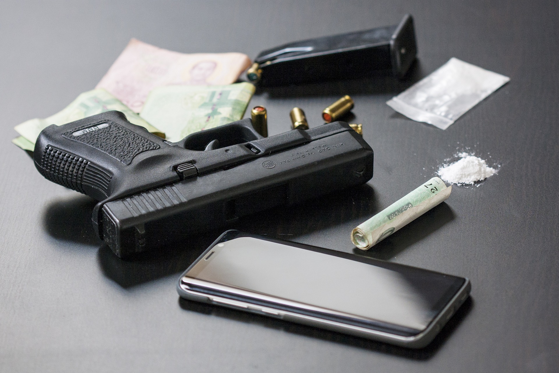 auf einer glatten schwarzen Oberfläche liegen eine Pistole, ein Smartphone und verschiedene Rauschgifte