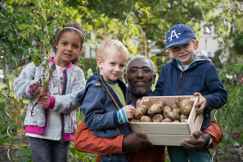 drei Kinder und ein Mann halten einen Kasten mit Kartoffeln und eine Gemüsepflanze hoch