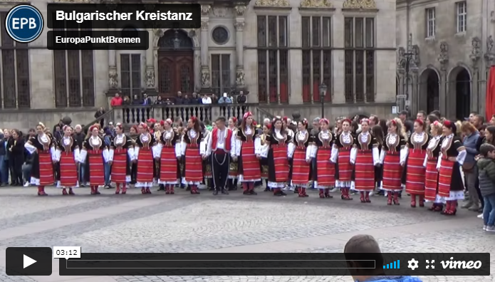 Eine Gruppe von Menschen tanzt im Halbkreis auf dem Bremer Marktplatz. Sie tragen Trachten. Im Hintergrund stehen Zuschauende.