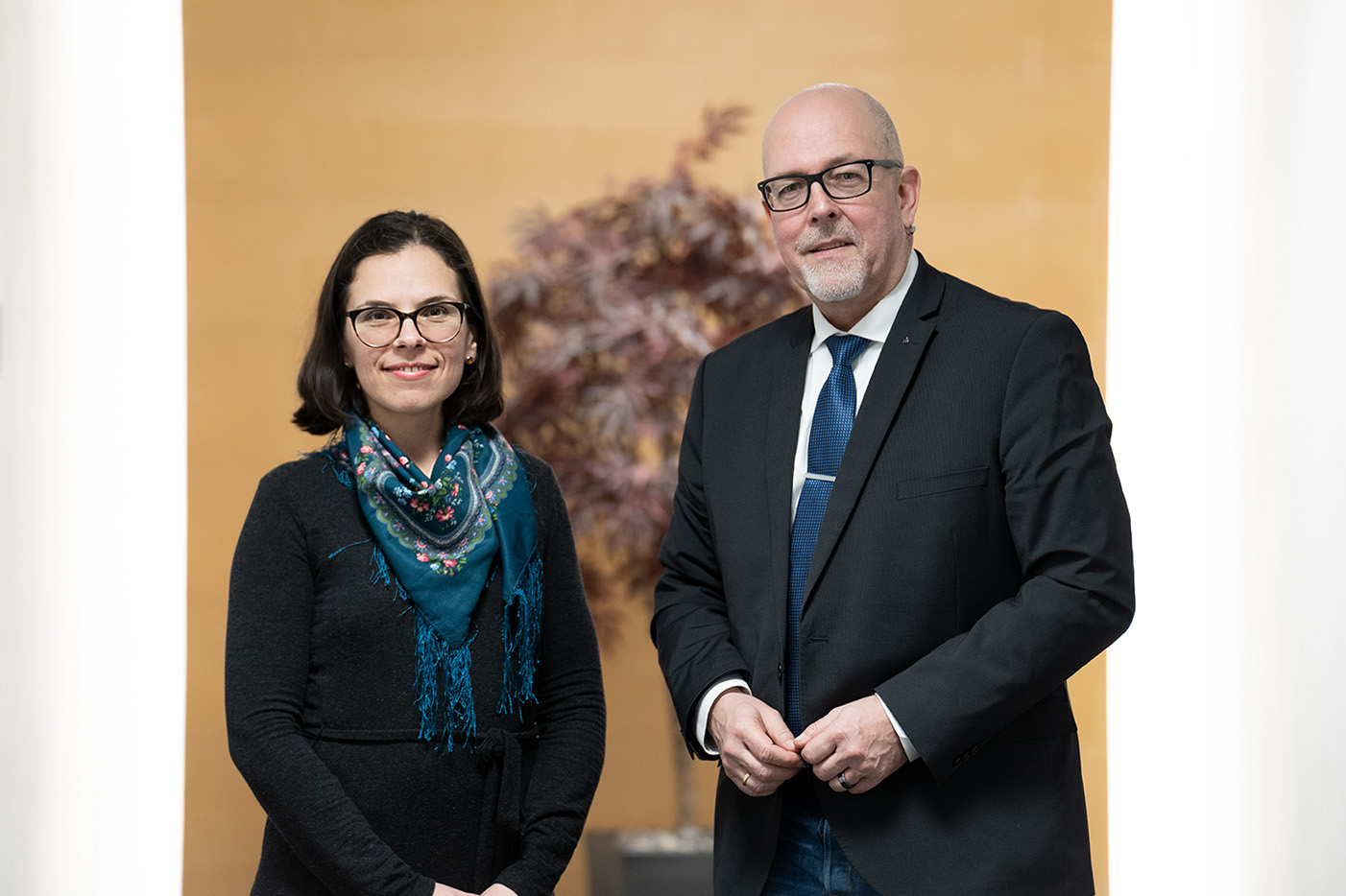 Eine Frau mit einer Brille und einem blauen Tuch um den Hals steht neben einem Mann in einem Anzug.