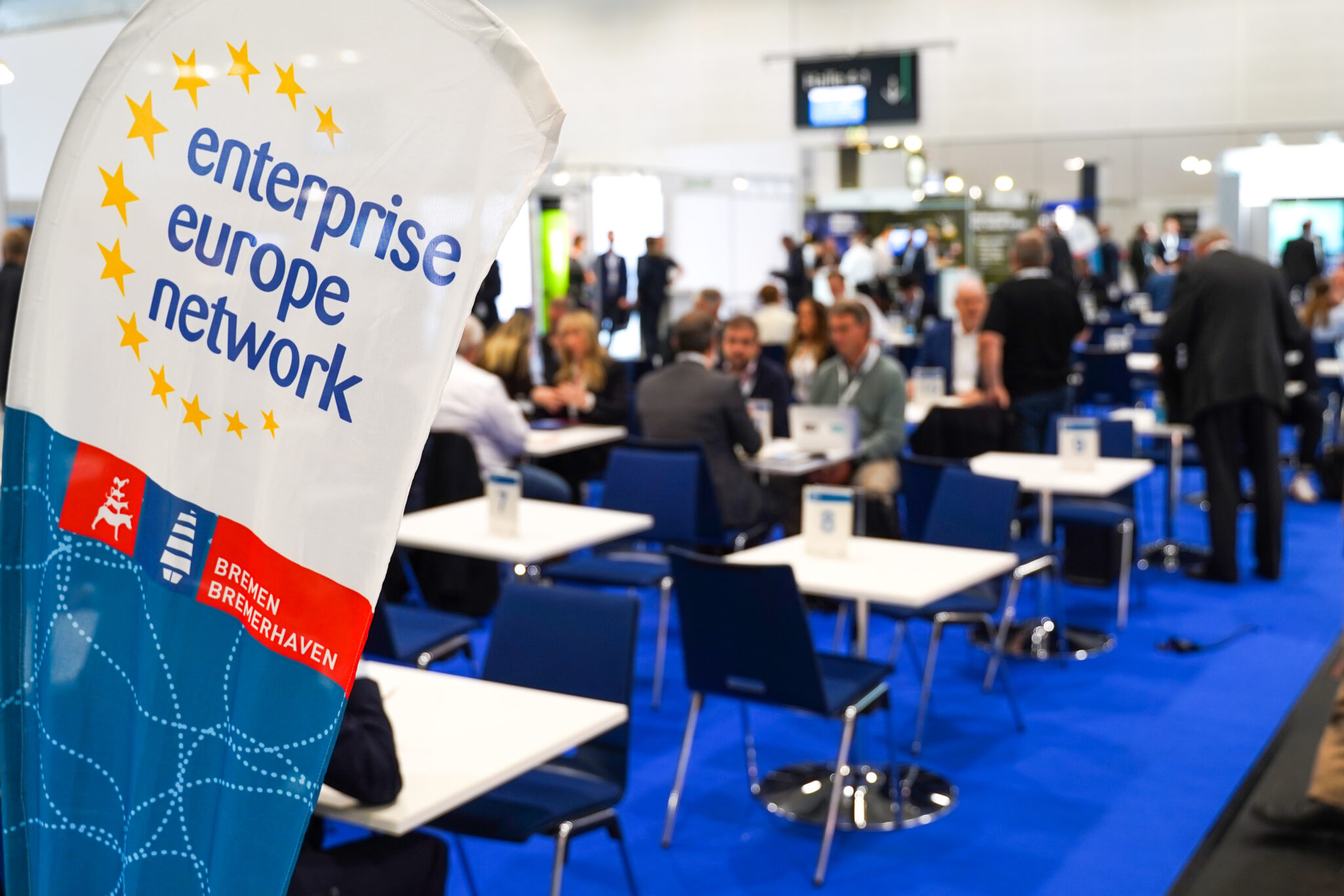 Im Fokus der Kamera eine Fahne mit der Aufschrift: enterprise europe network Im Hintergrund stehen mehrere Tische an denen ein paar Leute sitzen.