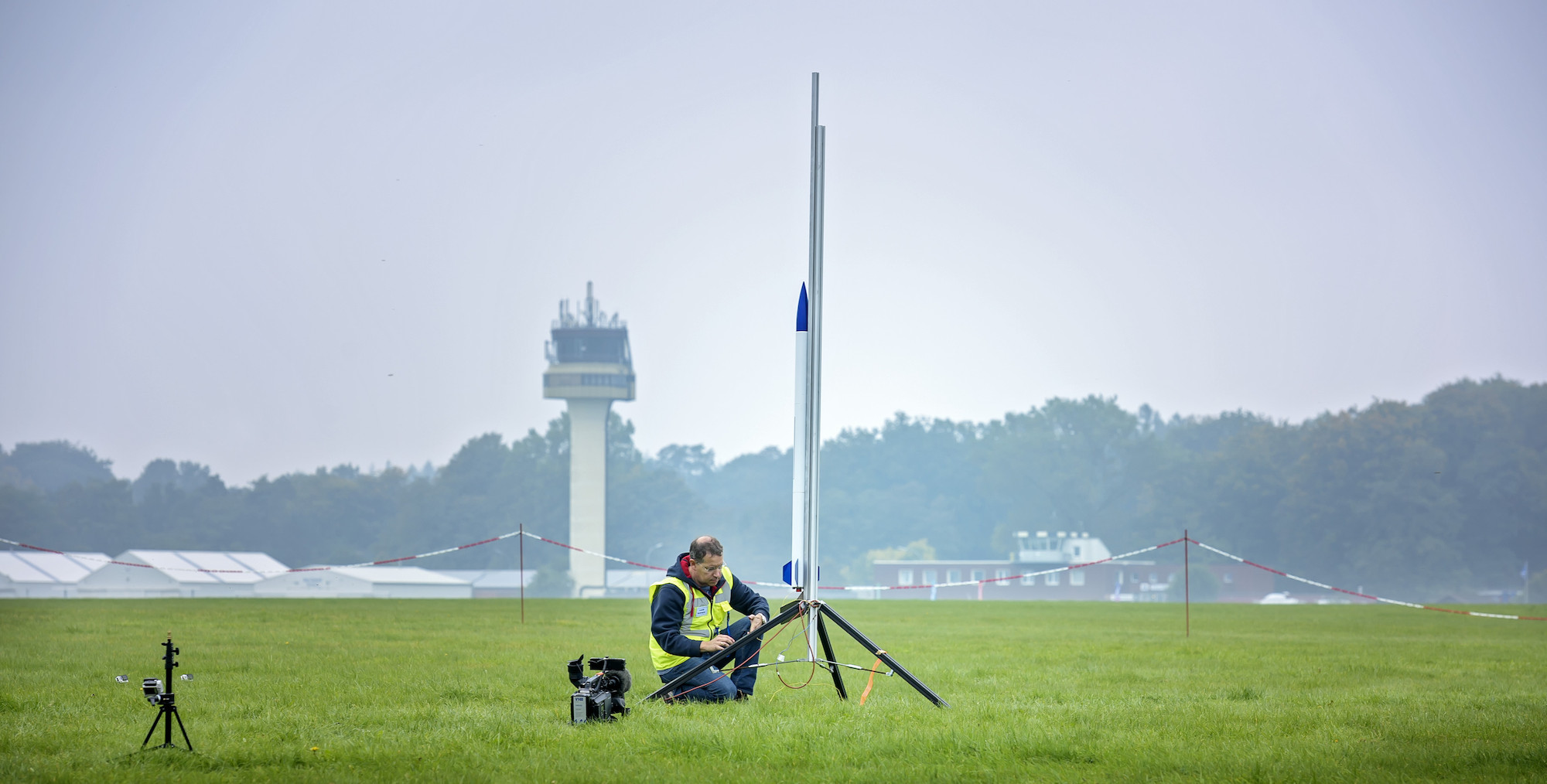 Ein Mann schraubt auf einer Wiese an einer kleinen Rakete, die an einem Gestell befestigt ist