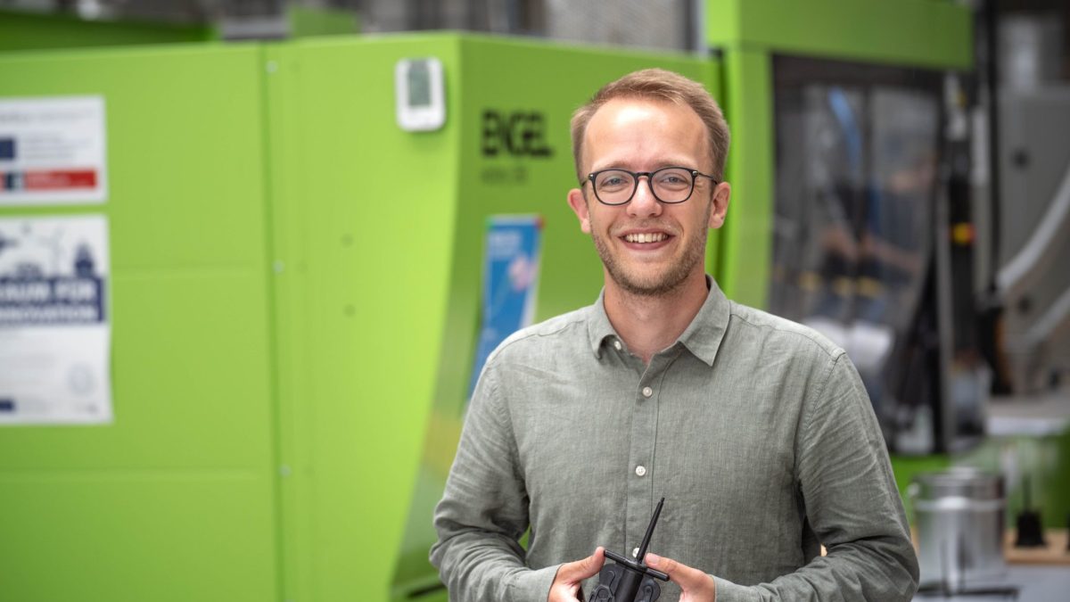 Ein Mann hält ein Kunststoffteil in der Hand und steht vor einer grünen Maschine