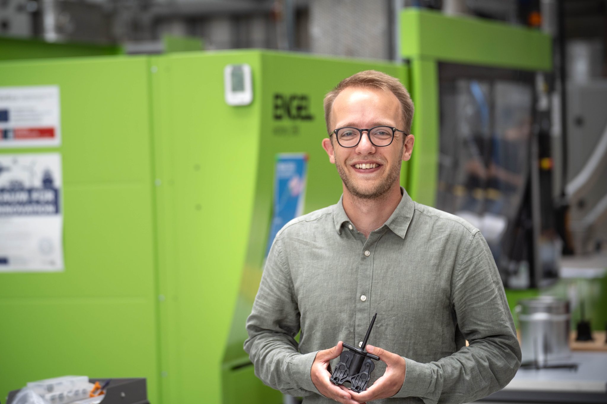 Ein Mann hält ein Kunststoffteil in der Hand und steht vor einer grünen Maschine