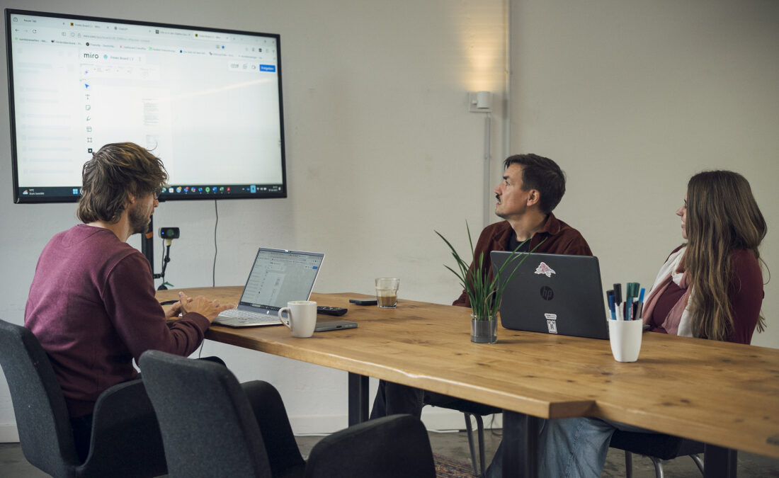 Bei einem Meeting von vomhörensehen sitzen drei Menschen an einem Tisch und schauen auf einen Bildschirm