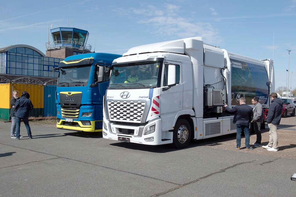 Ein weißer Hyundai-Brennstoffzellen-Lkw und ein blauer MAN-Lkw sind nebeneinander auf einer gepflasterten Fläche geparkt. Mehrere Personen stehen in der Nähe und unterhalten sich. Im Hintergrund sind ein Gebäude und ein Kontrollturm unter blauem Himmel zu sehen.
