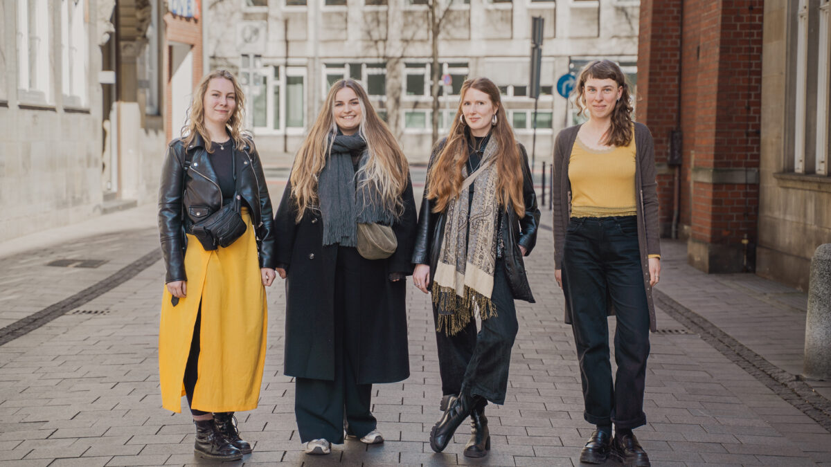 Vier Frauen stehen Seite an Seite auf einer Straße in der Stadt. Sie sind in Winterkleidung gekleidet, wobei zwei Frauen gelbe Röcke und die anderen dunkle Hosen tragen. Sie haben alle lange Haare und schauen leicht lächelnd in die Kamera.