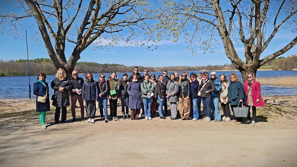 An einem sonnigen Tag posiert eine Gruppe von Menschen gemeinsam unter zwei großen Bäumen an einem See, im Hintergrund sind der blaue Himmel und das Grün der Natur zu sehen.