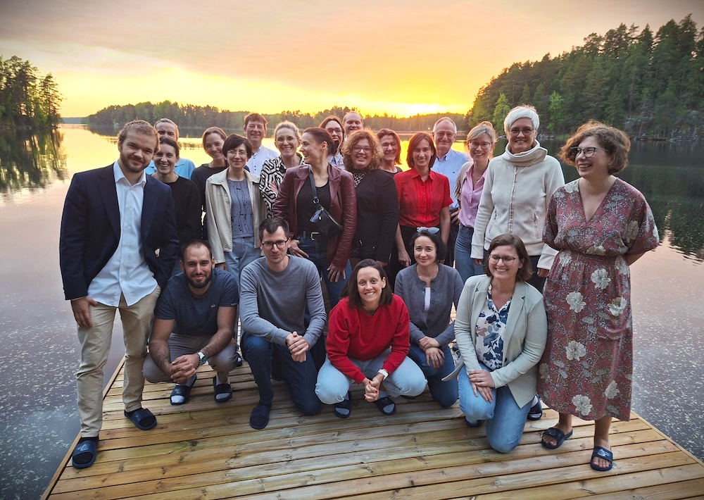 Eine Gruppe von etwa 20 Personen posiert gemeinsam bei Sonnenuntergang auf einem Holzsteg an einem See, umgeben von Bäumen. Einige stehen, andere knien, und alle lächeln und genießen die Naturkulisse.